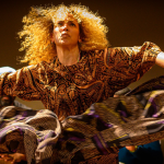 Image: Close up image of a woman dancing. She is dancing and looking towards the sky, in the direction of the camera. The woman's curly blond hair moves in the air along with her dance movement. She is wearing a brown, yellow and black paisley patterned shirt and a large black skirt.