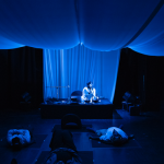 Image: Fabric is draped from the ceiling and the whole room is bathed in deep blue lighting. A Black woman sits on a stage with a microphone in her hand, surrounded by tank drums, electronics and a laptop. In front of her are four people lying down on rounded cushions on a black floor, spaced out six feet apart. By the entrance of the of the room, an attendant sits on a stool.
