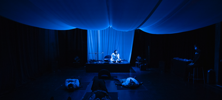 Image: Fabric is draped from the ceiling and the whole room is bathed in deep blue lighting. A Black woman sits on a stage with a microphone in her hand, surrounded by tank drums, electronics and a laptop. In front of her are four people lying down on rounded cushions on a black floor, spaced out six feet apart. By the entrance of the of the room, an attendant sits on a stool.