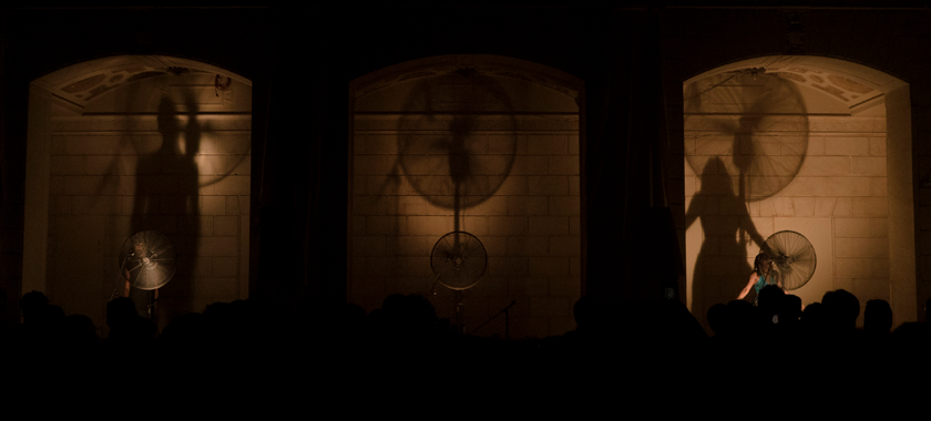 Image: Three industrial fans sit between three archways, sectioned off by massive pillars. The room is dimly lit in a golden light and shadows of the fans are projected on the walls behind them. In the far right and far left openings stand a Black man and a Black woman with microphone stands singing into the fans. The audience is seen in shadow, watching the performance unfold.