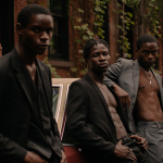 Image: Detail of a photograph of five individuals wearing blazers (some shirtless underneath) and dress pants, leaning against a maroon-colored vintage car; the image features earthy moody tones and various shades of Black skin.