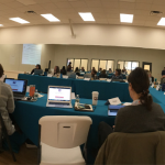 Image: Fisheye lens view of a professional development workshop at Arts Mid-Hudson; participants are seated at tables with paperwork and laptops as an individual stands at the front of a room with a presentation screen behind them