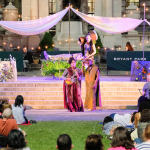 Image: View from the audience of a performance at Bryant Park; actors appear in a non-traditional stage setting as part of "Bryant Park Shakespeare" as the crowd enjoys from the grass and chairs.