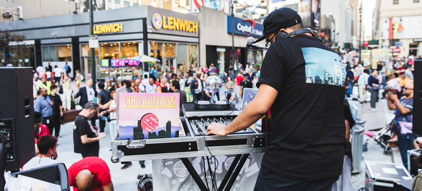 Image: A DJ wearing a black tee shirt, pants, and a hat performs at Plaza33 with turntables and electronic equipment; a record called "CHICAGOTRAX" is propped up with their setup.