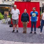 Image: Small outdoor improv comedy performance; four individuals stand together in a row, with two other individuals at the outskirts.