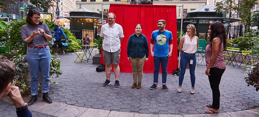 Image: Small outdoor improv comedy performance; four individuals stand together in a row, with two other individuals at the outskirts.