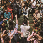 Image: A close-up photo of a crowd gathering at Bryant Park; audience members clap and dance and several individuals towards the center of the crowd hold instruments.