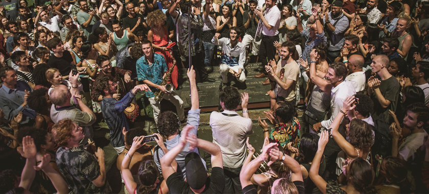 Image: A close-up photo of a crowd gathering at Bryant Park; audience members clap and dance and several individuals towards the center of the crowd hold instruments.