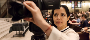 Photo of Luz Zamora looking into the small screen of a camera while adjusting the focus lens.