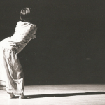 Image: Black and white photo of a solo performer on a stage; they are under a bright spotlight, and are pictured from behind in a dramatic pose.
