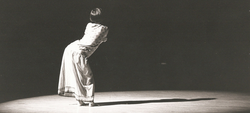 Image: Black and white photo of a solo performer on a stage; they are under a bright spotlight, and are pictured from behind in a dramatic pose.