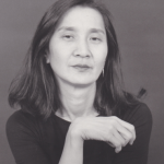 Image: Black and white portrait of choreographer Donna Uchizono. She has slightly below shoulder length dark hair and wears a crew-neck black tee-shirt and silver hoop earrings.
