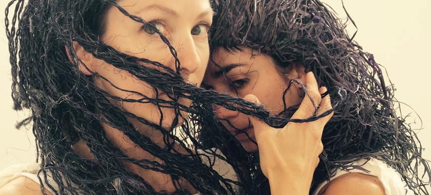 Image: Two performers photographed close up, one with face turned but eyes directly at the viewer, hand cradling the face of another performer who is less visible. Both wear wigs that appear to be made of straw.