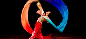 Image: a dancer is photographed against a black background, pictured in motion with a rainbow-colored long silk ribbon.