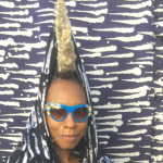 Image: Detail of a photograph of Nigerian-British musician Wunmi, who almost disappears into a blue and white patterned background. She is pictured from the shoulders up, wearing an outfit of the same design, hair braided and standing up straight on her head. She also wears bright blue sunglasses and a nose ring.