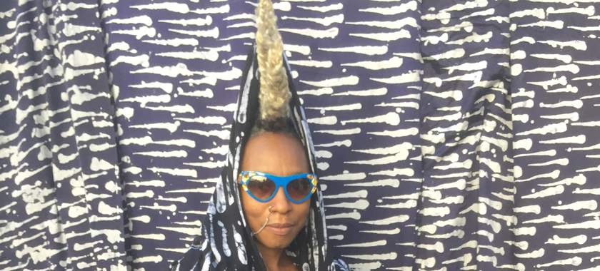 Image: Detail of a photograph of Nigerian-British musician Wunmi, who almost disappears into a blue and white patterned background. She is pictured from the shoulders up, wearing an outfit of the same design, hair braided and standing up straight on her head. She also wears bright blue sunglasses and a nose ring.