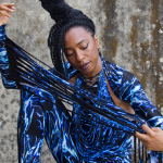 Image: Detail of a photograph of Nigerian-British musician Wunmi, pictured wearing a black leotard with blue and white water pattern and extra-long fringe on the arms that Wunmi pulls taut across the frame.