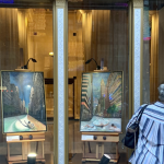 Image: two individuals are photographed from the back, looking onto glass store windows displaying two painted city scenes on easels. At the far left, there is Art on the Avenue signage in one of the store windows.