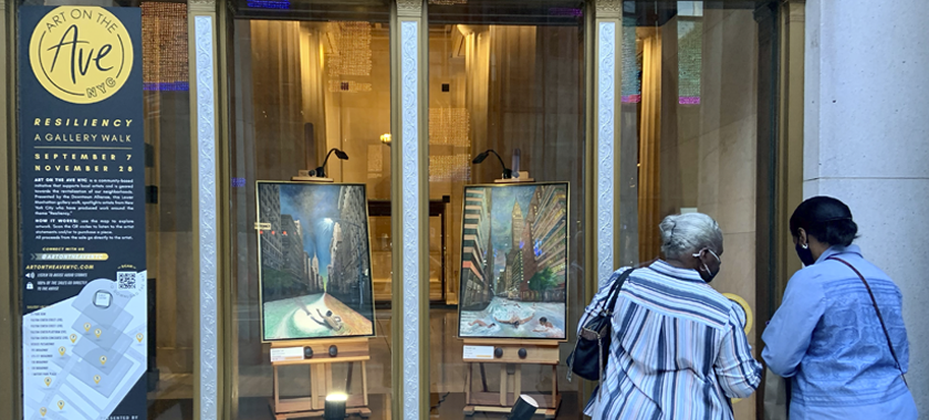 Image: two individuals are photographed from the back, looking onto glass store windows displaying two painted city scenes on easels. At the far left, there is Art on the Avenue signage in one of the store windows.