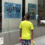 Image: Two individuals stand on a city sidewalk, looking into glass store windows at an Art on the Avenue display of colorful artworks.