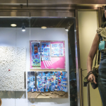Image: an individual is pictured on the right-hand side of the image, looking up and smiling. They stand in front of a store window with Art on the Avenue signage and art displayed behind a glass store window.