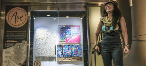 Image: an individual is pictured on the right-hand side of the image, looking up and smiling. They stand in front of a store window with Art on the Avenue signage and art displayed behind a glass store window.