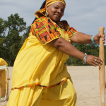 Image: An individual stands on a sandy beach smiling directly into the camera and wearing a bright yellow outfit. They hold a wooden instrument, with other instruments behind them in the background.