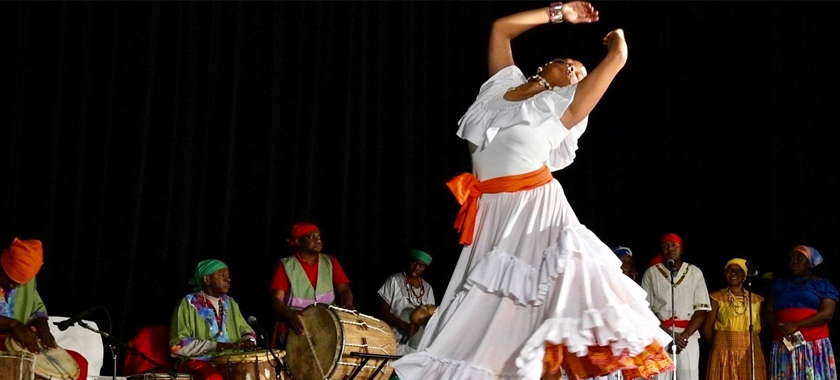 Image: A dancer in a white dress with orange accents leans back, arms above their head. In the background are singers and drummers accompanying the dance performance.