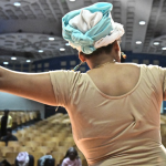 Image: Close-up of a dancer in rehearsal, photographed from the back with auditorium seating in front of them. They hold one arm out, hand up and rest their other hand on their hip.