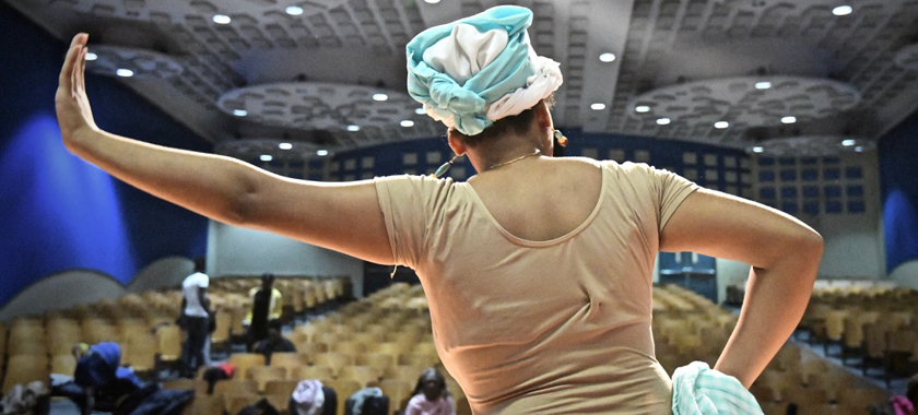 Image: Close-up of a dancer in rehearsal, photographed from the back with auditorium seating in front of them. They hold one arm out, hand up and rest their other hand on their hip.