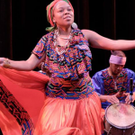 Image: A dancer is in the foreground, wearing a colorful printed outfit with arms outstretched. Individuals sit at drums in the background, providing music for the performance.