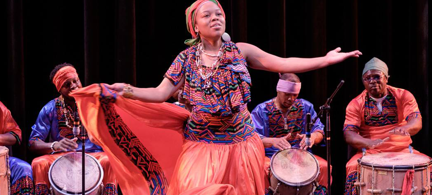 Image: A dancer is in the foreground, wearing a colorful printed outfit with arms outstretched. Individuals sit at drums in the background, providing music for the performance.
