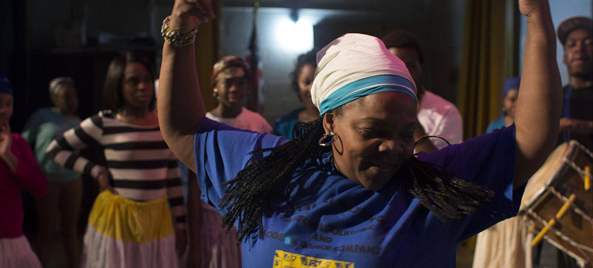 Image: An individual raises their arms above their head in movement as a group watches on in the background; their hair is long and braided, kept back with a headscarf.