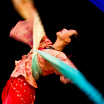 Image: a dancer is photographed against a black background, pictured in motion with a rainbow-colored long silk ribbon.