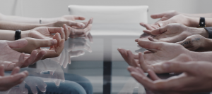 Image: Two lines of open-palmed hands mirror each other across a glass conference table. This is taken from a film called Drills, which is about the choreography of preparing for the future.