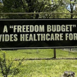 Image Detail: Artist Lizania Cruz standing in front of a large black banner with white writing that reads "A 'Freedom Budget' Provides Healthcare for All."
