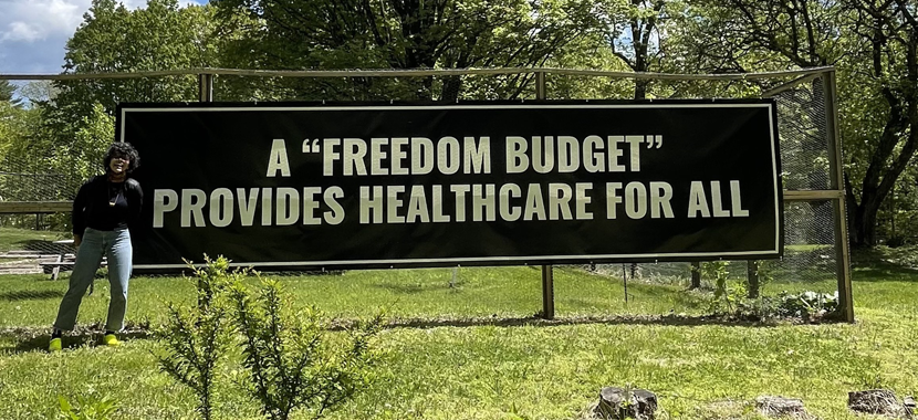 Image Detail: Artist Lizania Cruz standing in front of a large black banner with white writing that reads "A 'Freedom Budget' Provides Healthcare for All."