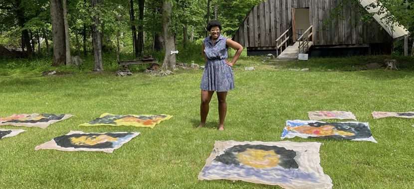 Image: Detail of a photo of artist Dana Robinson, who stands with one arm on her hip smiling into the camera. She stands on grass outside a wooden barn structure, her watercolor portraits on fabric in the grass around her.