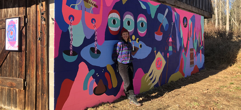 Image Detail: Artist Macon Reed leans against one side of a wood-roofed building with large wooden doors. On the wall where Reed leans is a brightly-colored surrealist mural design.