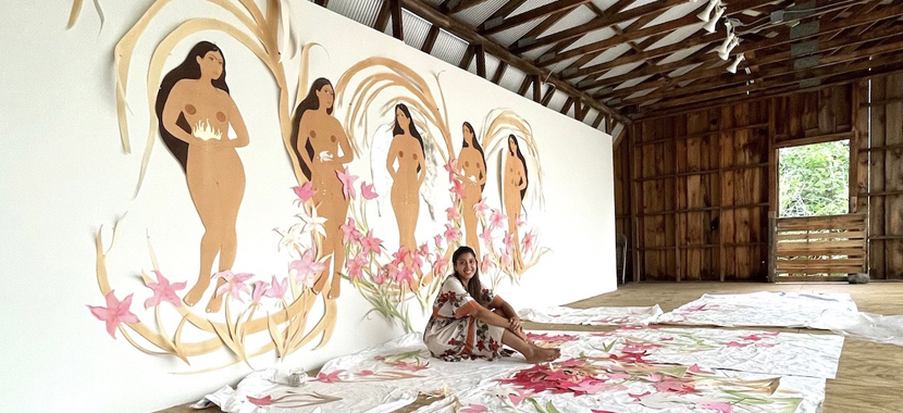 Image Detail: Artist Hiba Schahbaz in a large, barn-like structure with exposed beams. Her work appears on a mural behind her, and on fabric that is draped across the floor on which she sits. The mural features five paper cut-outs of nude women with flowing dark hair, swirls of paper and flowers around them.