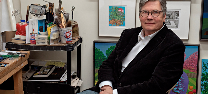 Image Detail: Photo of 2021 New Surrealist Prize recipient John Hrehov; he wears a white button down shirt, black velvet blazer, jeans, and glasses and looks directly into the camera. He sits with his artwork behind him and brushes and art supplies on a cart to his left.