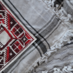 Image: variegated cotton embroidery thread on white Aida cloth, sewn onto a traditional Palestinian keffiyeh scarf, motif is approximately 6 x 6 inches. "The Goats" motif is a maternity motif because it features two goats facing one another, and in the white space between them, there are two baby goats. The artist embroidered this motif on the traditional Palestinian keffiyeh scarf, because it is a symbol of Palestinian identity to all in Palestine and in the diaspora. The scarf in totality represents a Palestinian mother -- a woman who loves to create and nurture.