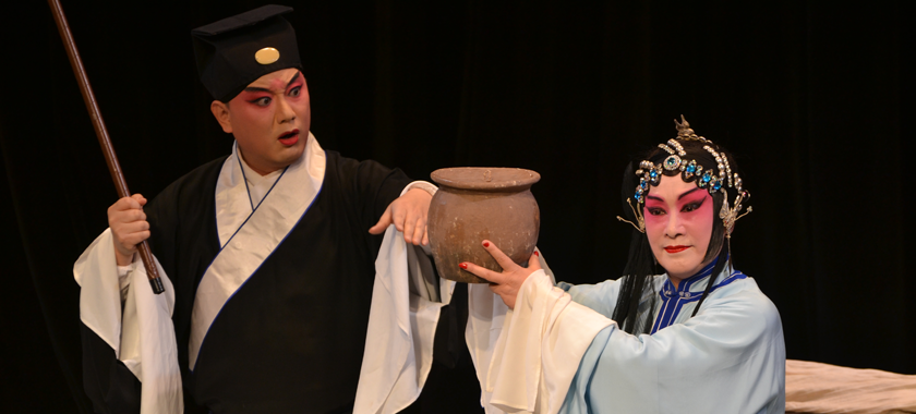 Image: Two individuals against a black background, lit by stage lights; they wear traditional dress and heavy makeup, one holding a bamboo pole and the other a clay pot.