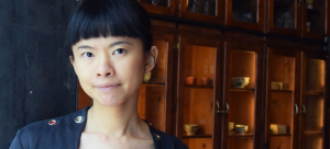 Image: Ya Yun Teng pictured against a floor to ceiling wood shelving display with ceramics inside; Teng has dark hair that is pulled back, bangs cut straight across her forehead. She wears a black top with grommets and looks directly into the camera.