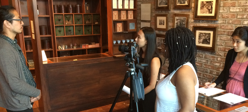 Image: Three interns interviewing an individual who is standing up and facing a camera. One intern appears to be interviewing the individual, another is behind the camera, and the third transcribing.