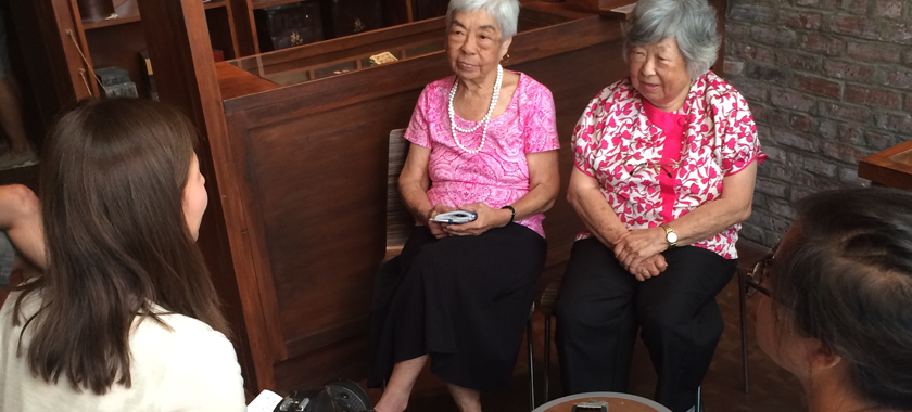 Image: Five individuals seated around a small table, the camera faces two elderly individuals in conversation with Museum of the Chinese in America interns.