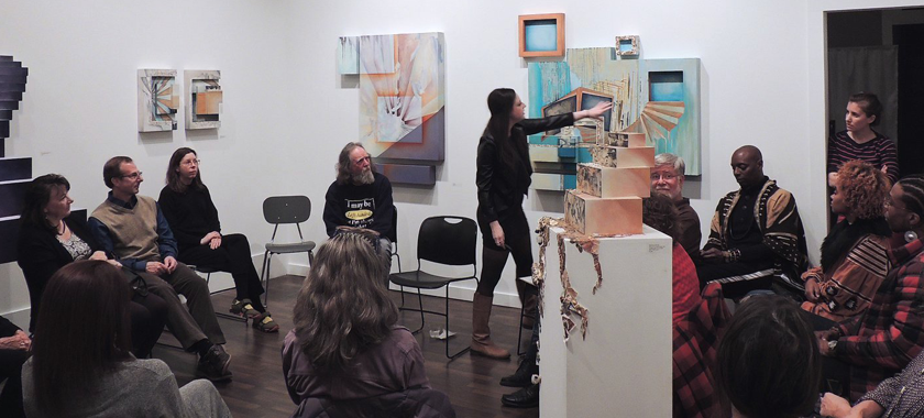 Image Detail: Kelly Olshan stands in the center of a photograph, gesturing towards a work that is hung on a white gallery wall and speaking to a seated crowd.
