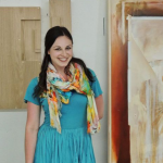 Image Detail: Kelly Olshan stands at the center of the frame, smiling and looking directly into the camera and wearing a turquoise colored dress. Her works are displayed behind her, with radiating lines and colors in earth and sky tones.