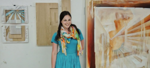 Image Detail: Kelly Olshan stands at the center of the frame, smiling and looking directly into the camera and wearing a turquoise colored dress. Her works are displayed behind her, with radiating lines and colors in earth and sky tones.