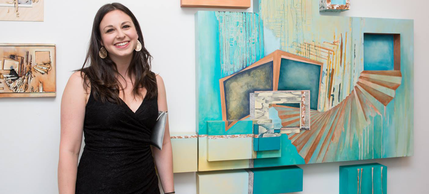 Image Detail: Kelly Olshan stands and smiles at the camera, dressed formally for an art opening of her work, which is displayed behind her on a white gallery wall.